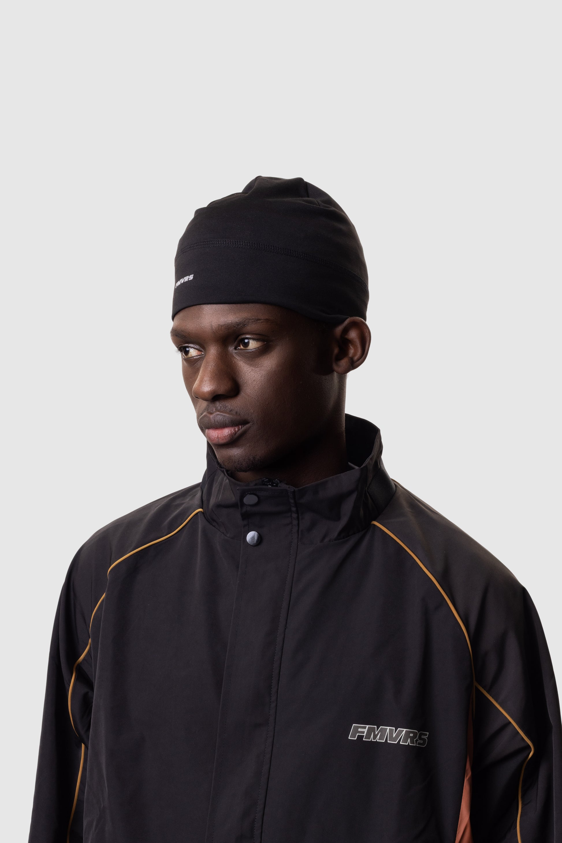 Man wearing a black jacket and beanie with 'FMVRS' branding on a light gray background